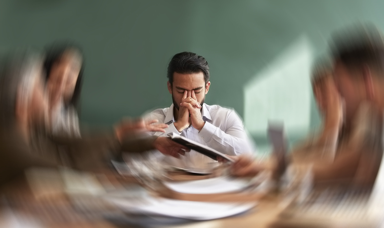 Homme épuisé en réunion, tête entre les mains, collègues flous autour de lui